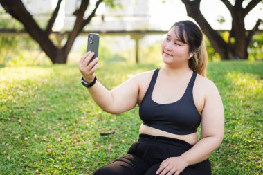 Bir kadının portresi çimenlerin üzerinde oturuyor, akıllı telefonuyla selfie çekerken gülümsüyor. Sahne rahattır ve doğadaki bir anlık keyfi yakalar. Asyalı artı beden kadın.