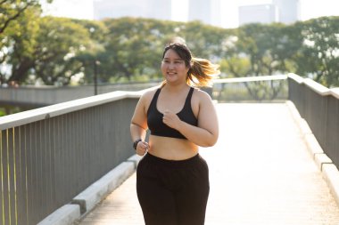 Neşeli bir kadının portresi, çevresini çevreleyen yeşillik, vücut bulmuş spor anlayışı ve aktif bir yaşam tarzıyla dolu bir köprüde koşuyor. Asyalı artı beden kadın.