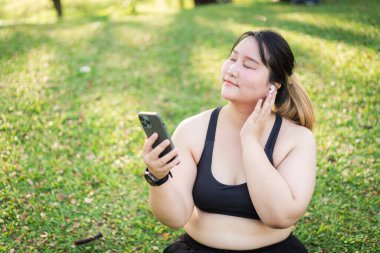 Bir kadının portresi açık havada, yeşil çimlerle çevrili telefonunu tutarken müzik dinlemekten hoşlanır. Asyalı ve cüsseli kadın sağlıklı yaşam tarzı, ilham verici görünüş, kendini sevme ve sağIık..