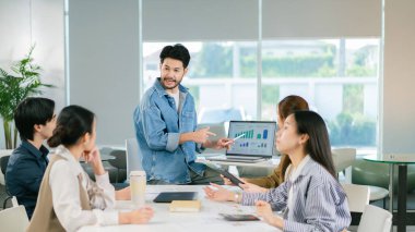 Bir adam modern ofis ortamındaki gruba iş verileri sunuyor, dizüstü bilgisayardaki grafikler ve şemalar sergileniyor. Atmosfer işbirlikçi ve profesyonel. Lider Takım Çalışması