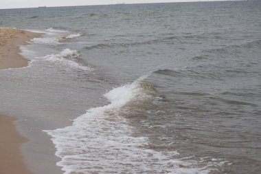 Fotoğraf kumlu bir kıyıya vuran deniz dalgalarını gösteriyor. Dalgalar ve kumla doğal kıyı manzarası