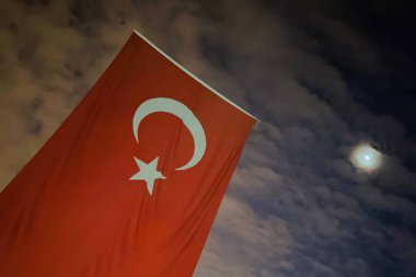 Ay Işığı Altındaki Türk Bayrağı. Hilal bir ay ve bayrağın üzerindeki sembolleri yansıtan yıldız ile gece dalgalanan Türk bayrağının çarpıcı görüntüsü. Doğal ay ışığı ve bulut deseni bu vatanseverlik sahnesine derinlik ve kontrastı ekler..