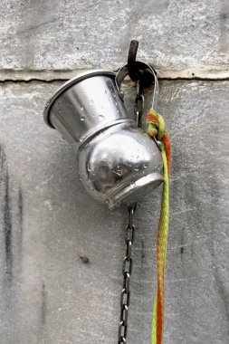 Hanging Metal Jug with Chain and Rope.The image shows a metal jug hanging on a wall. The jug is attached to a chain, which is secured to a hook on the wall. There is also a colorful rope tied to the chain.  clipart