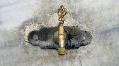 Taş Duvardaki Süslü Pirinç Musluk. Resim taştan bir duvara monte edilmiş süslü bir pirinçten musluğu gösteriyor. Musluk, biçimlendirilmiş ağaç ya da çiçek desenine benzer bir tasarıma sahip karmaşık ve dekoratif bir sapa sahiptir.. 