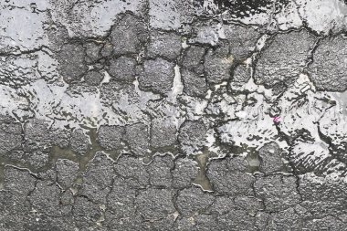 Islak Çatlak Asfalt Yüzey Aklama: Bu resim, ıslak ve çatlamış asfalt yüzeyin yakın çekimini gösteriyor. Asfalt yıpranmış ve yaşlanmış görünüyor, sayısız çatlak ve yamalarla.. 