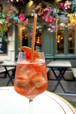 Paris Cafe Sett 'te Aperol Spritz. Canlı bir Aperol Spritz kokteyli, buzla parıldıyor ve portakal dilimiyle süsleniyor. Paris' te bir kafe masasında saplı bir bardakta zarif bir şekilde duruyor..