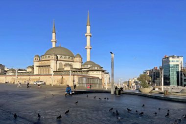 İstanbul, Türkiye. 2 Temmuz 2025 'te Taksim Meydanı ve Taksim Camii açık mavi gökyüzünün altında, güvercinler yerde toplanmış ve insanlar uzaklarda geziniyor. Sabah erken saatlerde Taksim Meydanı 'nda.