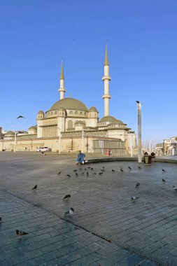 İstanbul, Türkiye. 2 Temmuz 2025 'te Taksim Meydanı ve Taksim Camii açık mavi gökyüzünün altında, güvercinler yerde toplanmış ve insanlar uzaklarda geziniyor. Sabah erken saatlerde Taksim Meydanı 'nda.