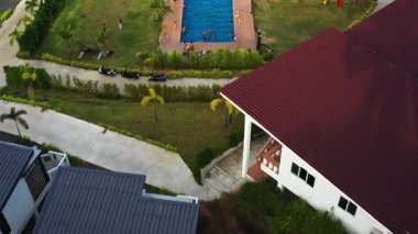 Çocuklar bir dinlenme tesisi havuzunda oynarken yetişkinler şezlonglarda rahatlarken drone öne doğru uçuyor. Palmiye ağaçları, fayans döşemeleri ve yakınlardaki bu huzurlu evde tüm sahneyi yakındaki evlerle yakalıyor.