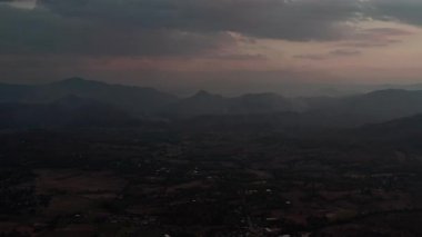 Alacakaranlıkta sakin bir dağ manzarası, dumanlı tepeler ve ön planda kırsal yerleşimlerle. Engebeli zirveler, alacakaranlıktaki gökyüzünün pembeden maviliğe dönüşmesine karşı dramatik siluetler yaratır.