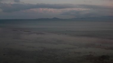 Renkli gökyüzü sakin sulara yansıyan panoramik günbatımı deniz manzarası. Güneş kısmen bulutlar tarafından gizlenmiş. Uzak adaların üzerine sıcak bir parıltı saçıyor. Islak kum desenleri ve yumuşak dalgalarla sakin kıyı manzarası