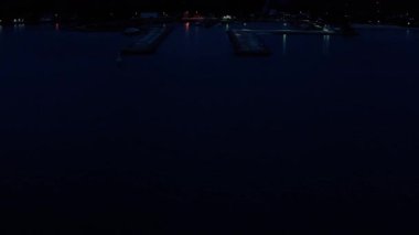 Sakin bir gece limanı, rıhtımlı tekneleri ve sakin karanlık suları yansıtan yapay ışıklarla hafifçe aydınlatılan bir balıkçı köyünün insansız hava aracı görüntüleri. Sahne sessiz bir güzelliği vurgular.