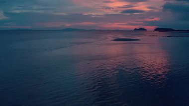 Alacakaranlıkta sakin bir sahil, yumuşak maviler, gökyüzünde mor ve portakallar, sakin sulara yansıyor. Kumlu sahil şeridi, uzak ada siluetleriyle dalgalanma desenleri gösteriyor. Dağınık bulutlar oluştur