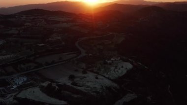 Güneş dağların üzerinden batarken, sakin bir günbatımı gökyüzünü turuncu, pembe ve mora boyar. Altın ışık, yuvarlanan tepeler ve dolambaçlı bir yoldur. Dağınık yapılar buna insan varlığını da katar.