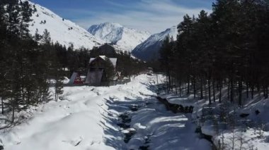 Kuzey Kafkasya 'daki Baksan Nehri geçidi üzerindeki İHA görüntüleri kış manzarasını gösteriyor. Karla kaplı ağaçlar, dik çatılı evler ve mavi gökyüzünün altında karla kaplı dağlar. Kısmen donmuş bir nehirle