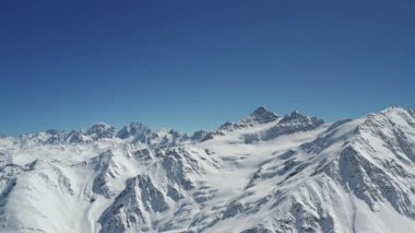 Elbrus Dağı 'nın panoramik manzarası ve açık mavi gökyüzünün altında karla kaplı tepeleri. Kış manzarasında pırıl pırıl kar ve engebeli tepeler vardır. Güneş ışığı bu dağdaki kardan yansıyor.