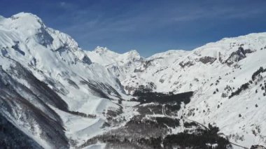 Elbrus Dağı bölgesindeki kış manzarasında engebeli kayalık yamaçlarla görkemli karla kaplı tepeler yer alıyor. Aşağıdaki huzurlu vadide koyu kozalaklı ağaçlar ve açık mavi bir kışın altında dolambaçlı bir yol var.