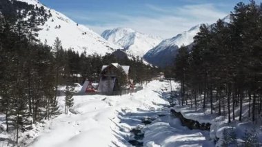Bir dron, karlı bir nehir vadisinde kırsal ahşap evleri ve üçgen bir kulübesi olan uçuyor. Çam ağaçları ve karla kaplı dağlar, berrak mavi bir gökyüzünün altındaki sahneyi çevreleyerek, huzurlu bir kış yaratıyor.