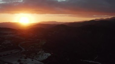 Sakin bir günbatımı, yuvarlanan tepeleri ve dağları altın ışıkla yıkıyor. Dolambaçlı bir yol, canlı turuncu, pembe ve mor renklerle boyanmış bir gökyüzünün altındaki tarlalardan geçer.