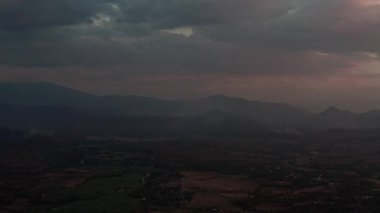 Pai 'nin dumanlı tepeleri dramatik bulutlu bir gökyüzü altında, engebeli dağ siluetleri ve altında bir vadi var. Dünyevi tonlar baskın, alacakaranlık ışıklarıyla güçlenirken, uzak dumanlar insana işaret ediyor.