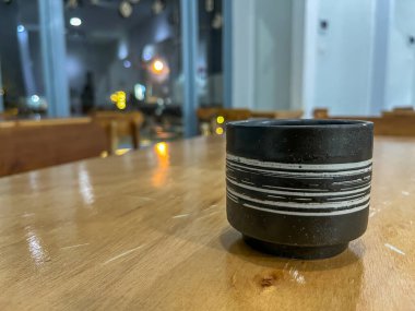 Ahşap masanın üzerinde beyaz çizgili siyah bir seramik bardak. Geceleri sıcak ışıklar ve bulanık bir arka planla rahat bir ortamda..