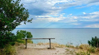 Ukrayna 'nın Cherkasy bölgesindeki Kaniv Reservoir' in sakin manzarası, bulutlu mavi gökyüzünün altındaki sakin sulara bakan basit ahşap bir bank..