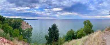 Ukrayna 'nın Cherkasy bölgesindeki sakin Kaniv Reservoir' in panoramik bir görüntüsü. Uçurumlar ve parçalı bulutlu gökyüzünün altındaki yeşil ağaçlarla çevrili.