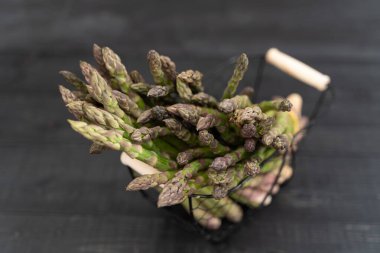 Metalik sepetin içine yerleştirilmiş yemyeşil kuşkonmaz, hasattan hemen sonra, siyah ahşap arka planda.