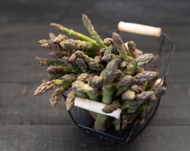 Üzerinde siyah ahşap bir masanın üstüne yerleştirilmiş metal bir sepete yerleştirilmiş yeşil kuşkonmaz resmi var..