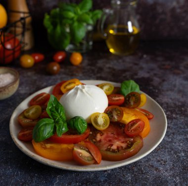 Ortasında burrata olan karışık domatesler, fesleğen yaprakları, zeytinyağı ve koyu mavi kahverengi arka planda bir kase deniz tuzu.