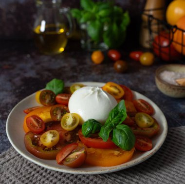 Dilimlenmiş domatesli burrata, fesleğen yaprağı, zeytinyağı ve koyu mavi kahverengi arka planda bir kase deniz tuzu.