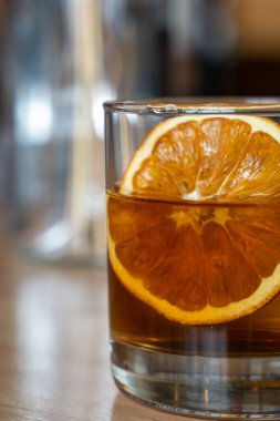 Viski bazlı bir kokteyl, büyük kurumuş portakal dilimleriyle süslenmiş kısa bir bardakta servis edilir. İçki, yumuşak ortam aydınlatmasında ahşap tonlu bir masanın üzerinde duruyor..