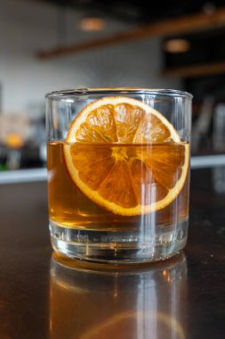 Kristal berrak bardakta sunulan koyu kehribar bir içecek, büyük bir portakal dilimi garnitür içeriyor. Huysuz ışıklandırmayla yansıtıcı bir barın tepesinde çekilmiş..
