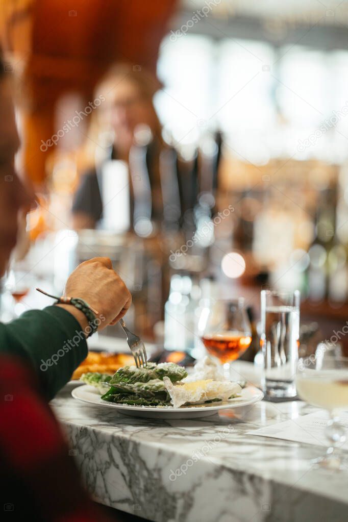 Person enjoying a Caesar salad and drinks at a marble bar with a lively restaurant atmosphere.