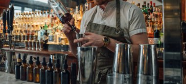 Barmen dikkatli bir teknikle bir kokteyl hazırlıyor. Parlak bar aletleri ve kaliteli içkilerle çevrili sofistike bir barda..