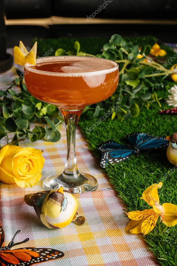 Close-up of a crown-garnished cocktail with a yellow rose and butterfly props on checkered fabric.