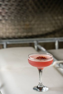 Pembe bir kokteyl, kurutulmuş kan portakal dilimiyle süslenmiş, yumuşak bir endüstriyel yüzeye sunulmuş bir coupe bardakta servis edilir..