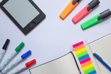 Masadaki Renkli Kırtasiye ve Dijital Okuma Aygıtı. Dijital okuma cihazı, renkli kalemler, bir defter, yapışkan notlar ve bir kalemin yer aldığı düzenli bir çalışma alanı. Verimliliği, organizasyonu ve öğrenim ortamlarını resmetmek için ideal