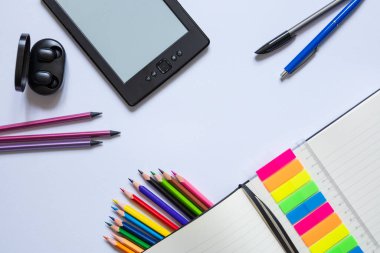 Not Defteri ve Gadget ile Yaratıcı Çalışma Alanı. Renkli kalemler, yapışkan notlar, e-okuyucu ve kulaklıklar beyaz çalışma alanı arka planında düzgünce düzenlenmiş.