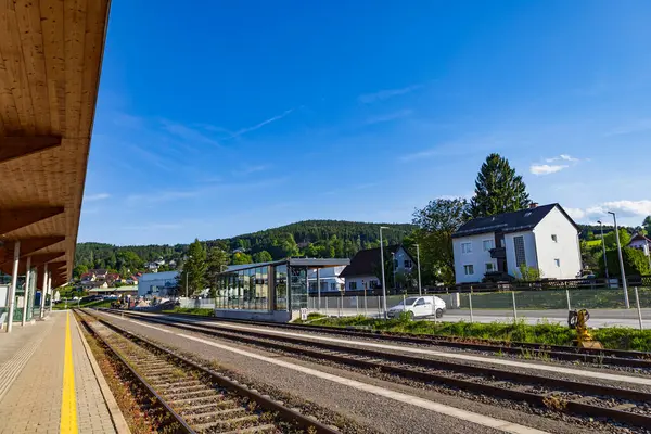 Tepelerde yuva yapmış küçük bir köye doğru uzanan rayları olan sakin bir tren istasyonu, modern mimari ve aydınlık, güneşli bir gün, kırsal yolculuğun güzelliğini vurguluyor.