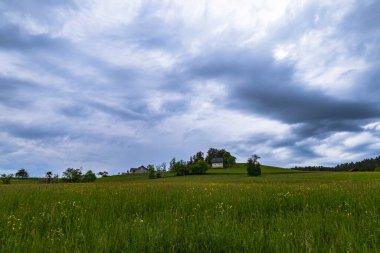 Huzurlu bir manzaraya yerleşmiş küçük bir kır evi, kasvetli bir gökyüzünün altında yeşil tarlaların arasında duruyor. Sakin kırsal bölge ile yaklaşan fırtına arasındaki zıtlık sahneye dinamik bir unsur ekliyor.