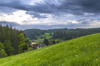 Yuvarlanan tepelerin ve yemyeşil yeşilliklerin arasına yerleşmiş sakin bir köy. Parçalı bulutlu bir gökyüzü altında büyüleyici evler ve canlı doğa sergiliyor..