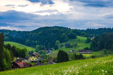 Yuvarlanan tepelerin ve yemyeşil yeşilliklerin arasına yerleşmiş sakin bir köy. Parçalı bulutlu bir gökyüzü altında büyüleyici evler ve canlı doğa sergiliyor..