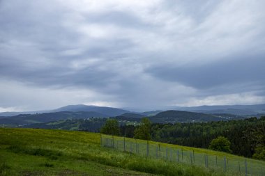 Çarpıcı, bulutlu bir gökyüzü altında yuvarlanan tepelerin ve yemyeşil çayırların manzarası. Manzarada bir çit ve dağınık ağaçlar var. Huzurlu ve sakin bir atmosfer yaratıyor.