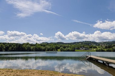 Bereketli yeşil tepeler ve berrak mavi gökyüzü ile çevrili ahşap iskele ile sakin bir göl manzarası.