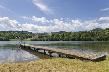 Tepeler ve Orman 'la Lakeside İskelesi. Bereketli tepeler ve ormanlarla çevrili sakin bir göle uzanan ahşap bir iskele.