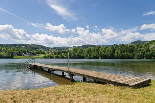Tepeler ve Orman 'la Lakeside İskelesi. Bereketli tepeler ve ormanlarla çevrili sakin bir göle uzanan ahşap bir iskele.