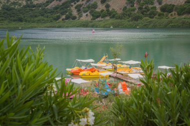Georgioupoli, Chania, Girit. 4 Haziran 2025. Zakkum çiçeklerinin arasından Kournas Gölü manzarası. Tornavidalı bir adam katamaranların yanındaki tahta bir köprüde duruyor. Yüksek kalite fotoğraf