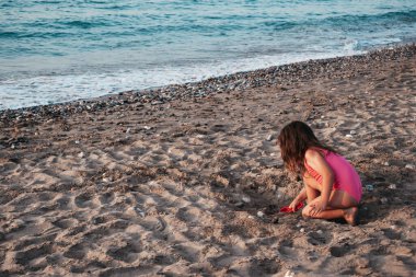 Pembe mayolu küçük bir kız kumsalda oynuyor, kumda kırmızı bir kürekle çukur kazıyor. Yüksek kalite fotoğraf