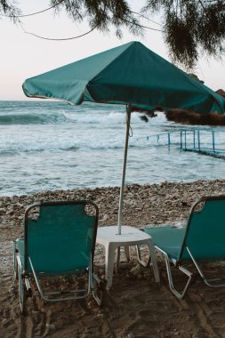 Gün batımında plaj sahnesi turkuaz şemsiyeli ve güneş şezlonglu, şezlonglar arasında küçük masa, küçük dalgalı deniz. Yüksek kalite fotoğraf